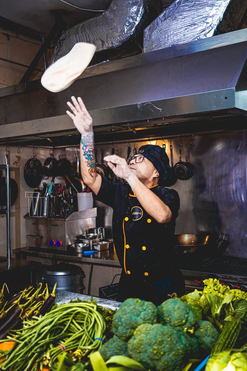 Chef tossing dough above a counter of fresh vegetables